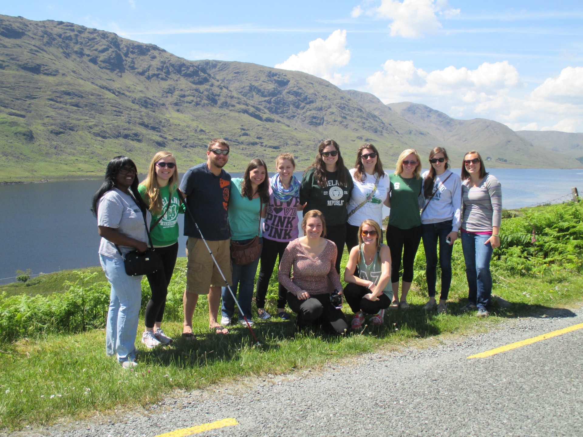 Study abroad students standing roadside in front of a lake and green mountain range 