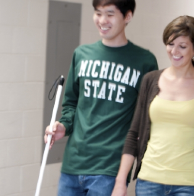 Two students walking together, one wearing a yellow shirt with a brown cardigan, and one wearing an MSU shirt and carrying a white cane.