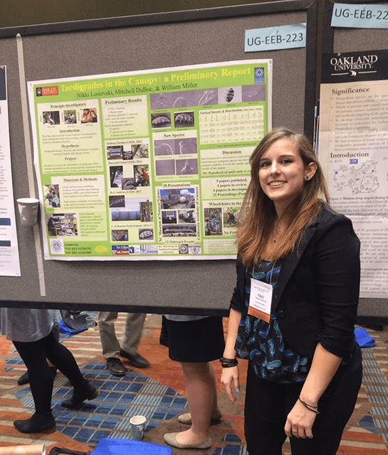 Photo of student smiling in front of presentation board at a conference.