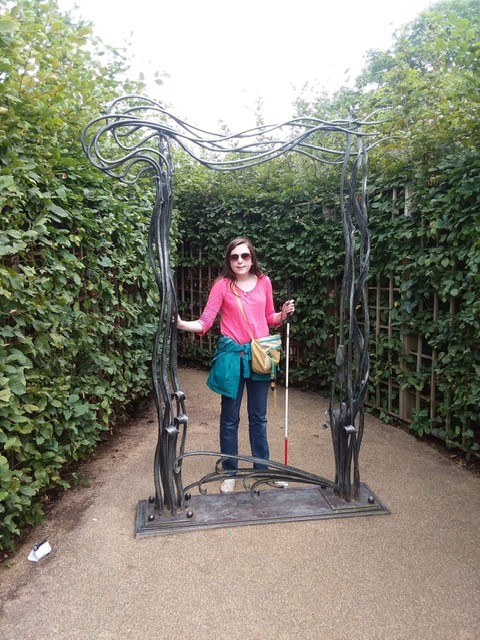 Photo of Ellie posing with her cane in hand, next to an ornate metal statue in a leafy garden