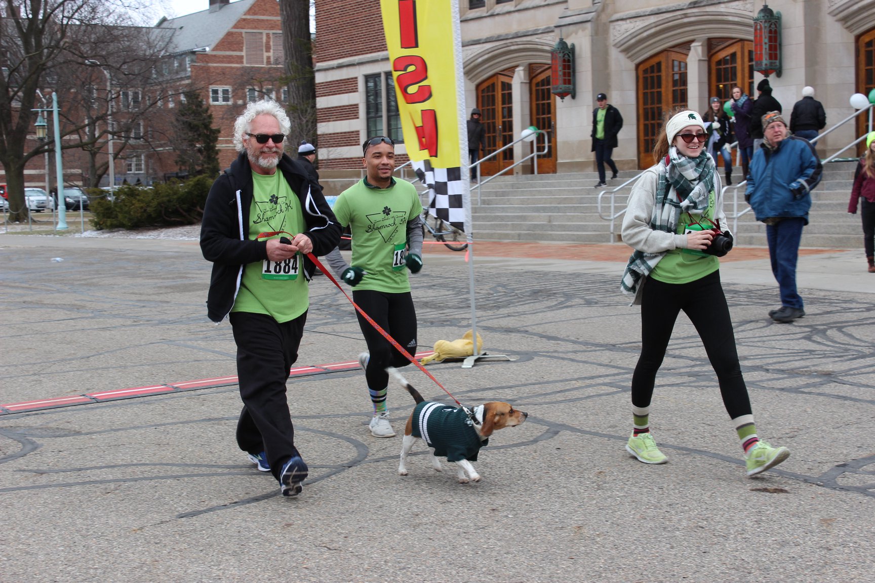 Three people walkind past the finish line. One is walking a beagle