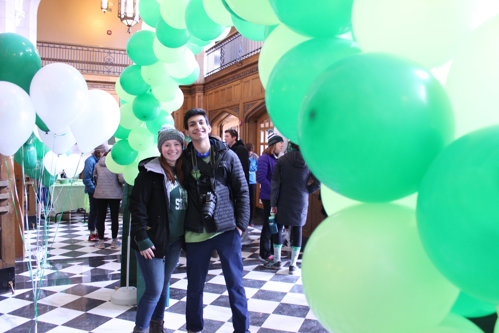 Two people smiling under an archway of green and white balloons