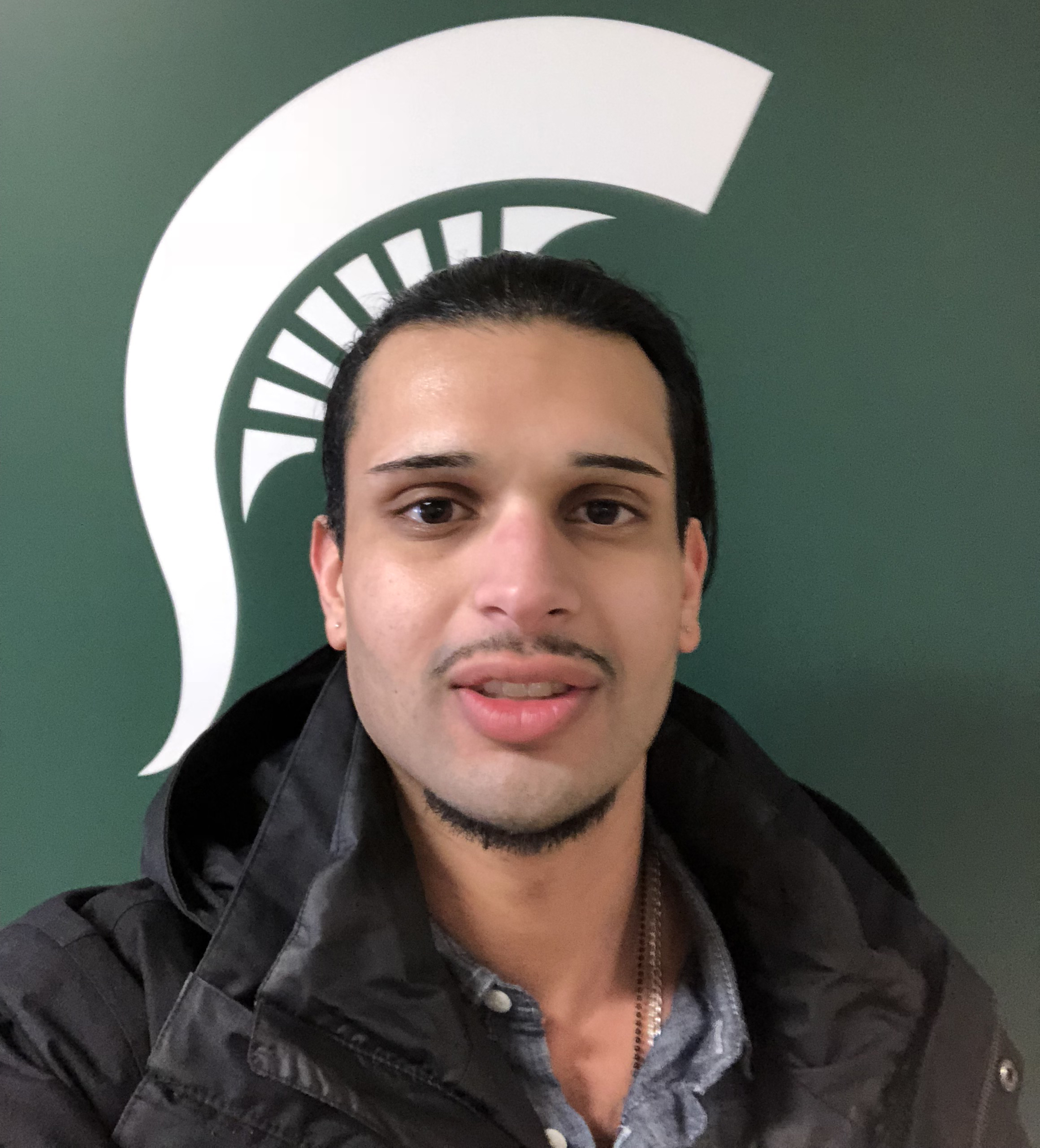 Photo of Neman H. standing in front of green and white Spartan Helmet logo