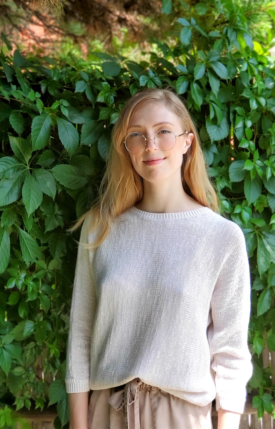 Elaine, grinning Caucasian woman, wears a white sweater and khaki pants, with glasses and light brown hair.