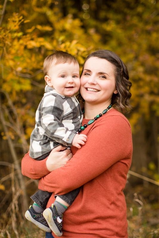 Liz wears orange sweater, holding her son, who wears black and white plaid shirt, grey pants, and black and white shoes.