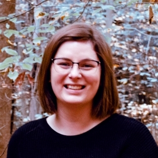 Pictured is Tesia, Causcaisian woman, with shoulder length brown hair and square glasses. She wears a black scoop neck top (or dress), with plants in the background.