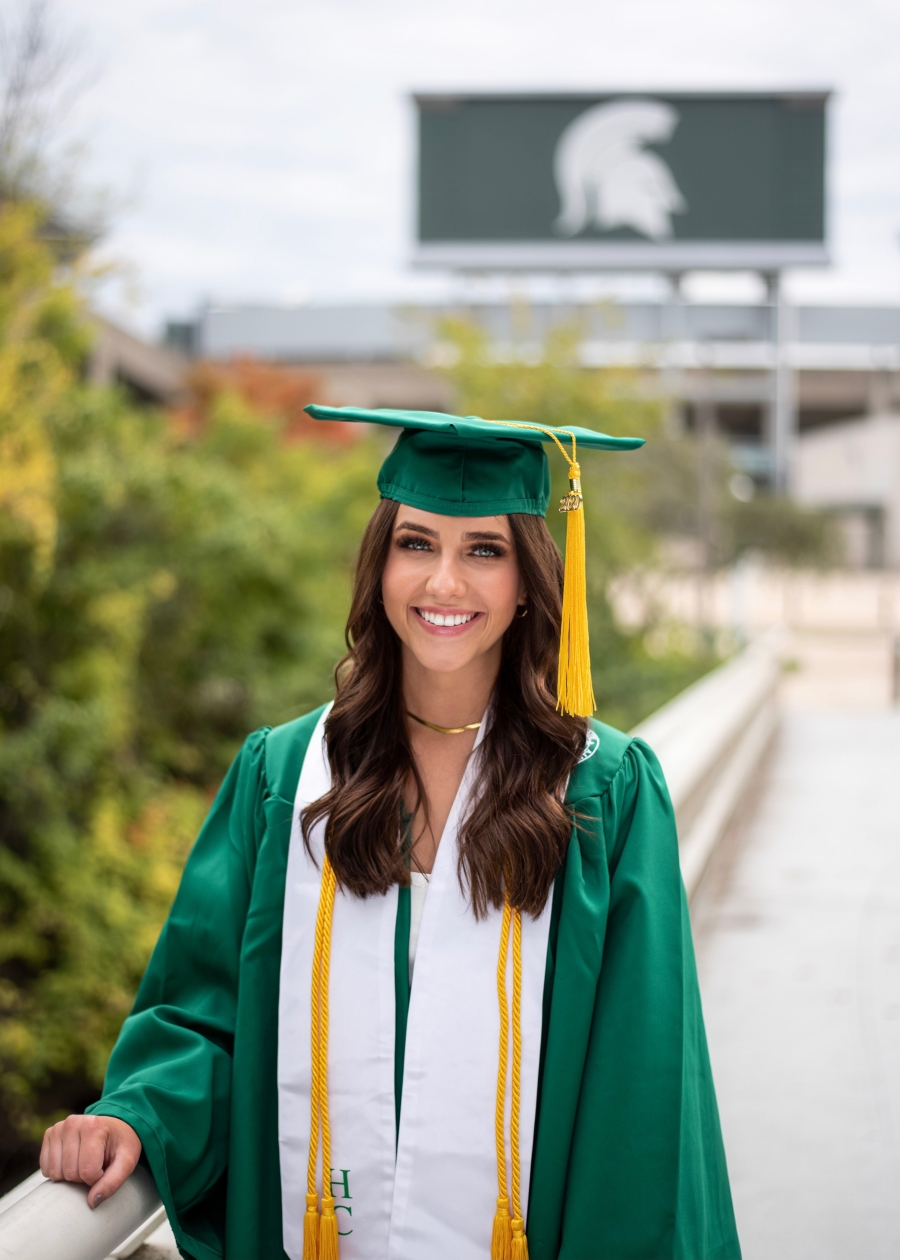 Pictured is Anna, wearing MSU cap and gown, smiling in front of the Spartan stadium. She wears long, wavy brown hair.