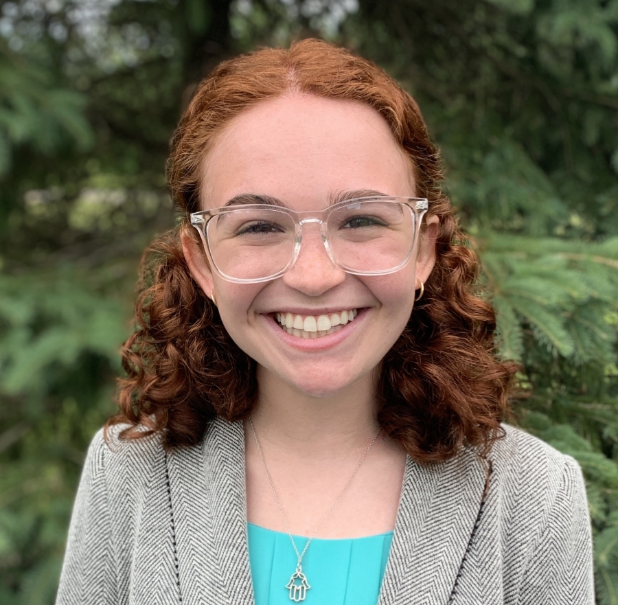 Pictured is Laya who wears a smile with curly red hair, clear square glasses, a teal blouse, and a grey blazer jacket. 