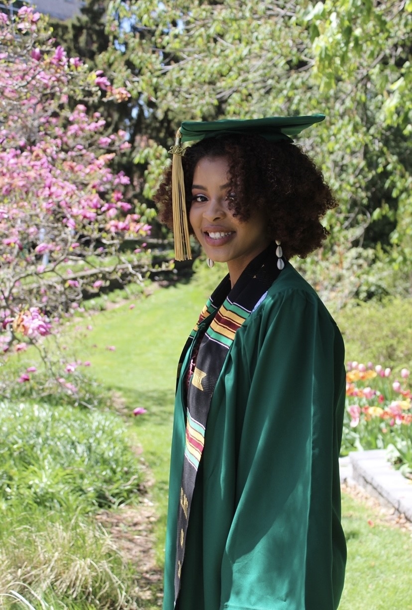 Picture of Jasmine J., smiling Black female, with curly dark brown hair, wearing green cap and gown and a black, African American Celebratory stole. She is pictured in front of green trees and pink flower.