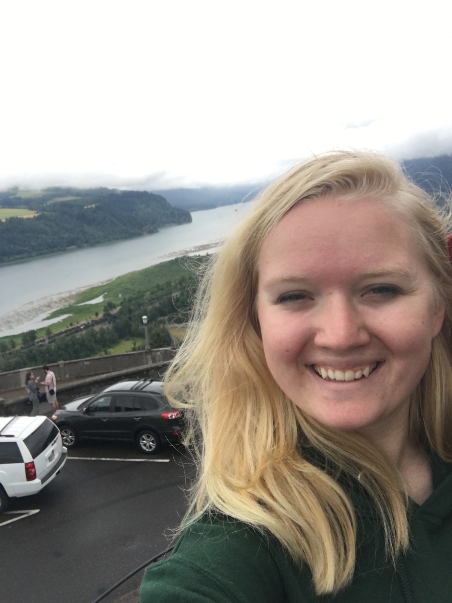 A photo of Emma, a young Caucasian woman with blond hair, with a hilly and foggy landscape behind her. 