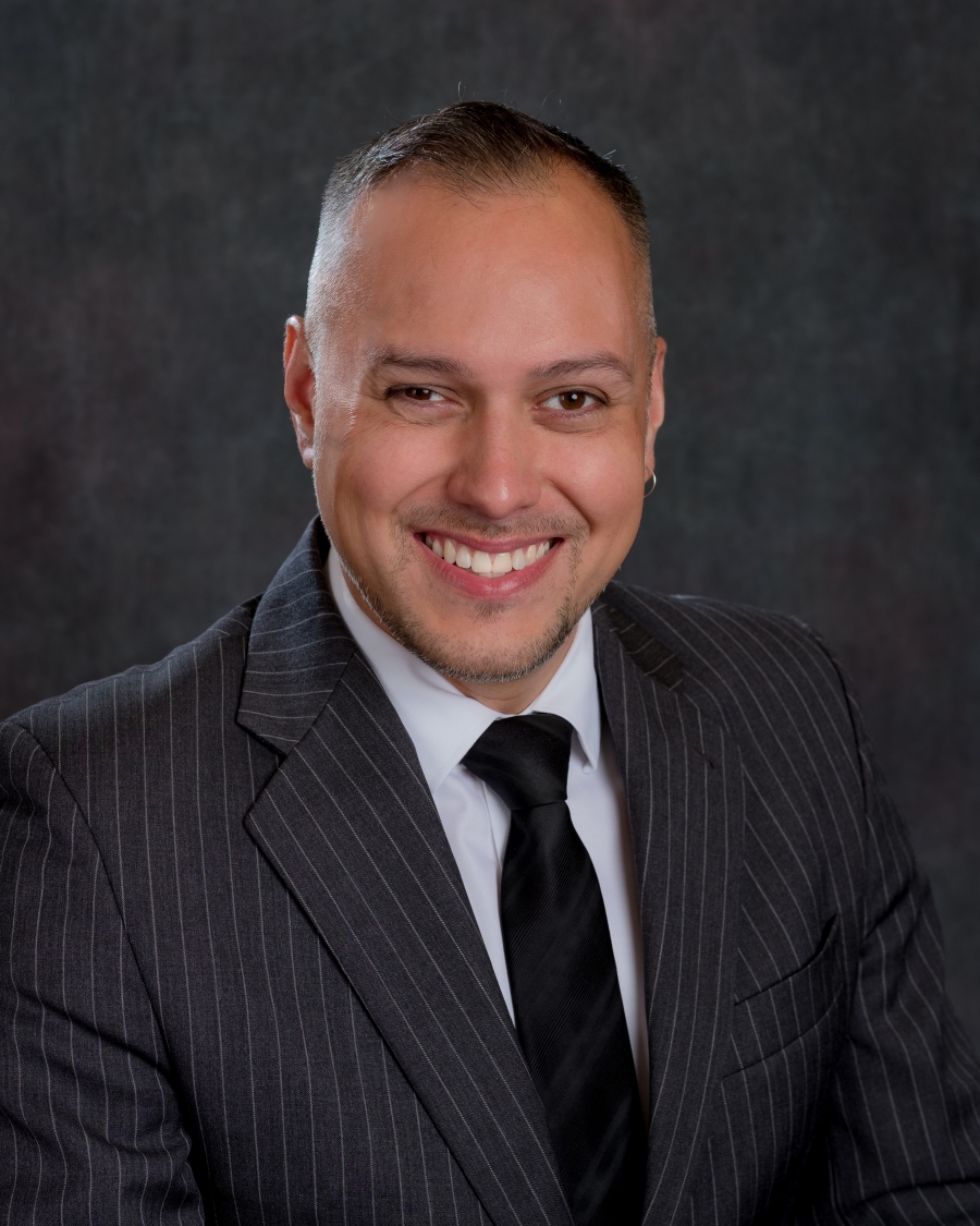 A headshot of Caleb. He his wearing a black and white pinstripe suit with a white shirt and black tie