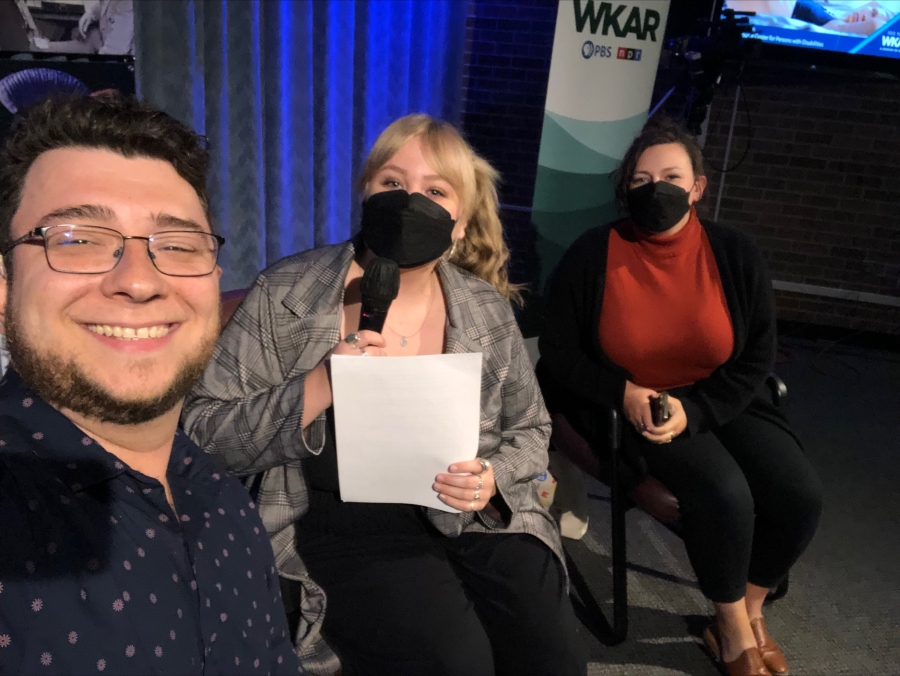Image of Tyler Smeltekop, Colleen Floyd, and Emily Abrams. Tyler is smiling and is wearing a black shirt. Colleen Floyd is wearing a mask and a gray blazer and is holding a paper and a microphone. Emily is wearing a mask and a red shirt and black cardigan  