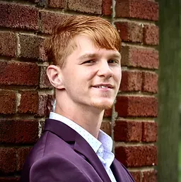 Headshot of Tyler Mazone. He has ginger hair and is leaning against a brick wall. He is wearing a purple blazer with a white dress shirt underneath.