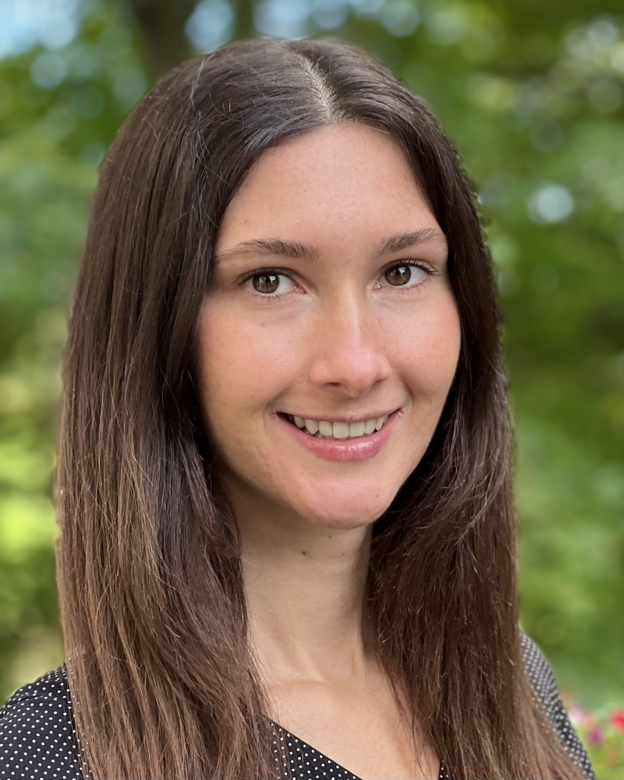 Headshot photo of Megan smiling; she has long, straight, brown hair parted down the middle and has greenery behind her.