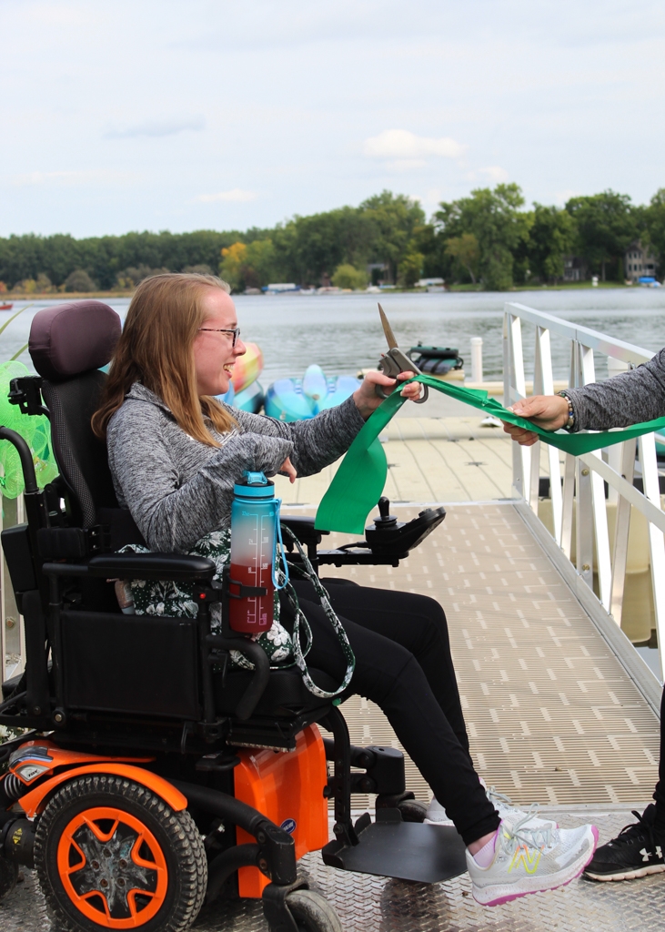 Photo of a woman in a motorized wheelchair cutting the green ribbon at the opening of the new accessible dock