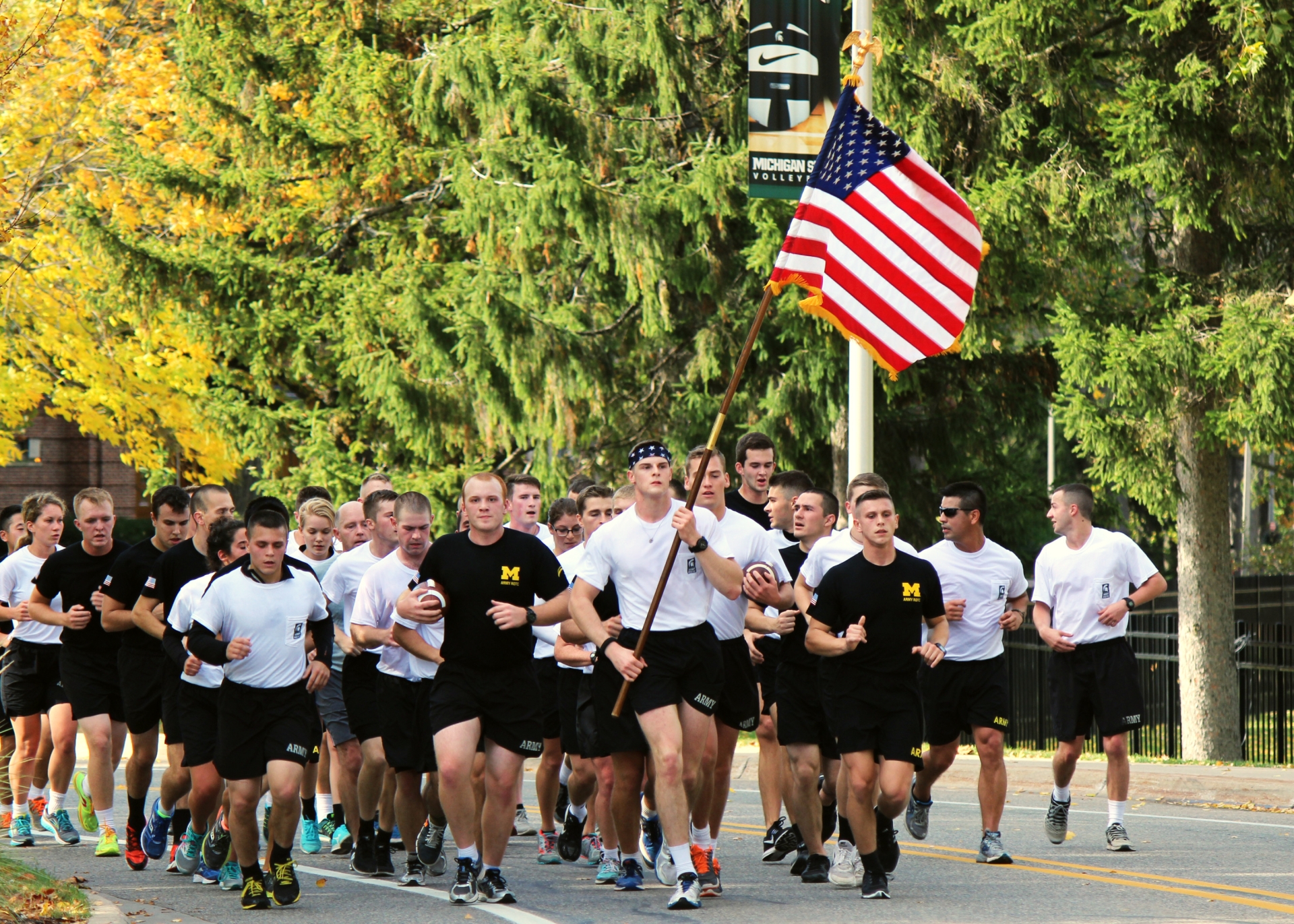 A group of ROTC cadets run down a street with trees in the background, holding a football and an American flag 