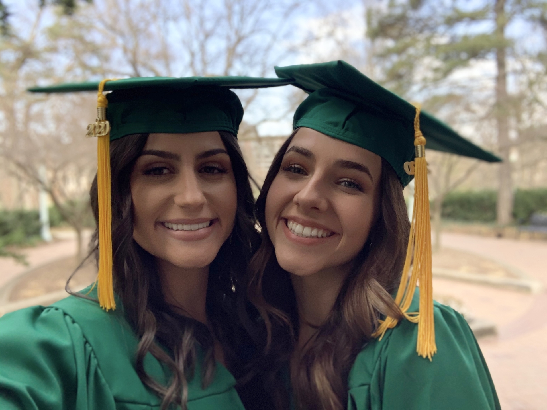 Pictured is Abby on the left and Anna on the right. They both wear MSU graduation caps and gowns, smiling with trees behind them.