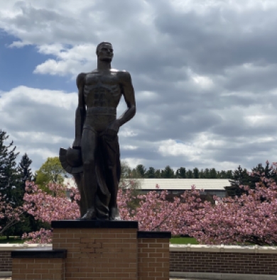 Pictured is the Spartan statue with a blue sky and clouds. In the background, there are pink flower and various shades of green trees.