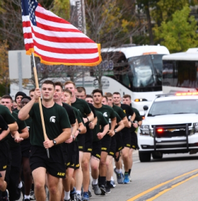 Army ROTC cadets running in the street with an American flag and a police escort