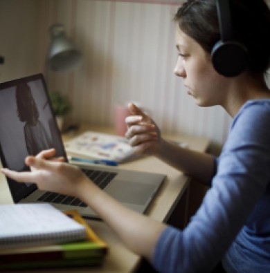 Pictured is a Caucasian woman wearing brown hair in ponytail, with black headphones on top, and a blue long sleeve shirt rolled up. She is on her laptop watching something with a notebook besides her.