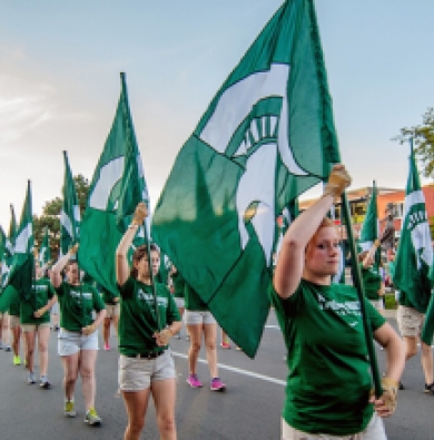 The Spartan color guard marching in a parade and hiding green flags with spartan helmets on them 