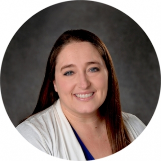 Photo of Shelby Gombosi - a smiling light skinned woman with long brown hair. She is wearing a blue V-neck blouse and a white sweater.