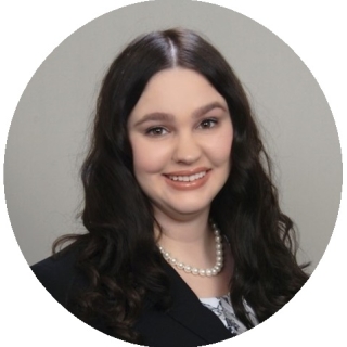 Photo of Kiersten Manos - A smiling Caucasian woman with long, wavy, black hair. She is wearing a black blazer over a white floral shirt with a pearl necklace.