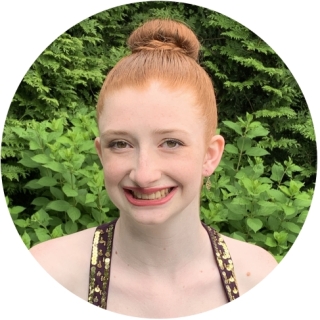 A photo of Julia Feuerborn -A smiling white woman with red hair in a bun. She is wearing a dark purple pleated top with gold sequins straps. She is wearing gold earrings.