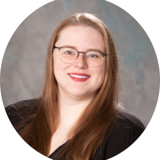 Photo of Ellie Brigger: A light skinned woman with blue eyes, freckles, and long, straight, dark blonde hair smiles at the camera. She is wearing round acrylic glasses and a black blouse with red, green, white and yellow flowers in front of a light grey background.