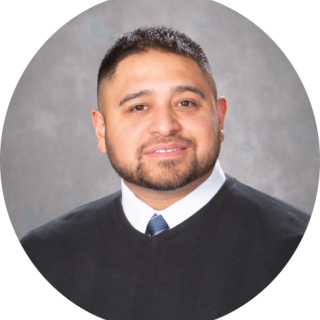 Photo of Robert Briseno, a man with short brown hair and a beard smiling at the camera, wearing a black sweater over a white button-up and tie.