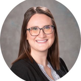 Photo of Kristin Campbell, a light skinned woman with collar-bone length brown hair smiling at the camera, wearing a black cardigan over a white shirt and she has glasses on