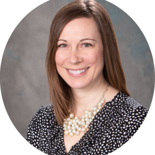 Photo of Rachel Giddings - a light skinned woman with collar-bone length brown hair smiling at the camera, wearing a black and white patterned shirt.