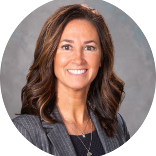 Photo of Suzie Hoffman, a light skinned woman with medium length brown hair smiling at the camera, wearing a gray blazer over a black shirt.