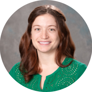 Photo of Hannah Huey-Jones- A smiling light skinned woman with wavy brown hair smiling at the camera, wearing a green shirt.