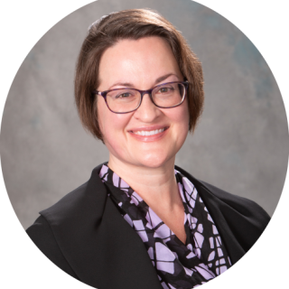 Photo of Virginia Rutan, a light skinned woman with short, bob cut brown hair smiling at the camera, wearing glasses and a patterned shirt under a black blazer