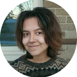 A person smiling with shoulder-length brown layered hair. They have brown eyes and tan skin and are wearing an earth-toned sweater with beige, brown, dark green, and red patterns. They have a septum piercing and a nostril piercing. The background of the picture shows brown bricks and a window. 